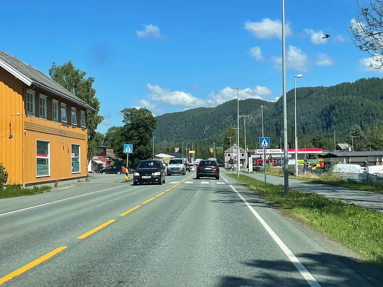 Lundamo sentrum sett fra E6 en sommerdag, med gult trehus, biler på vegen og bensinstasjon.