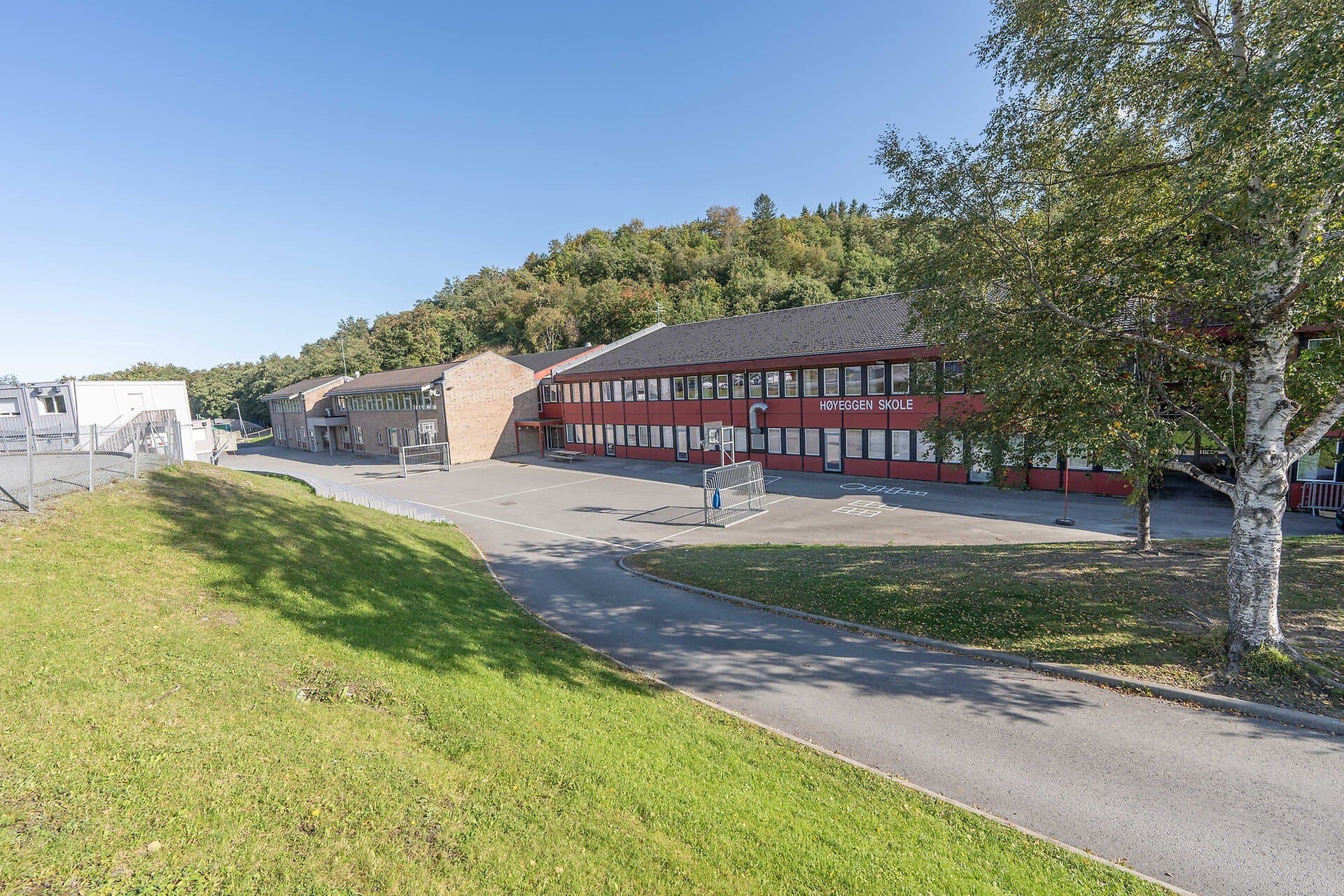 Høyeggen skole – rødt toetasjes skolebygg med skoleplass, gressplen og skogkledd ås bak.