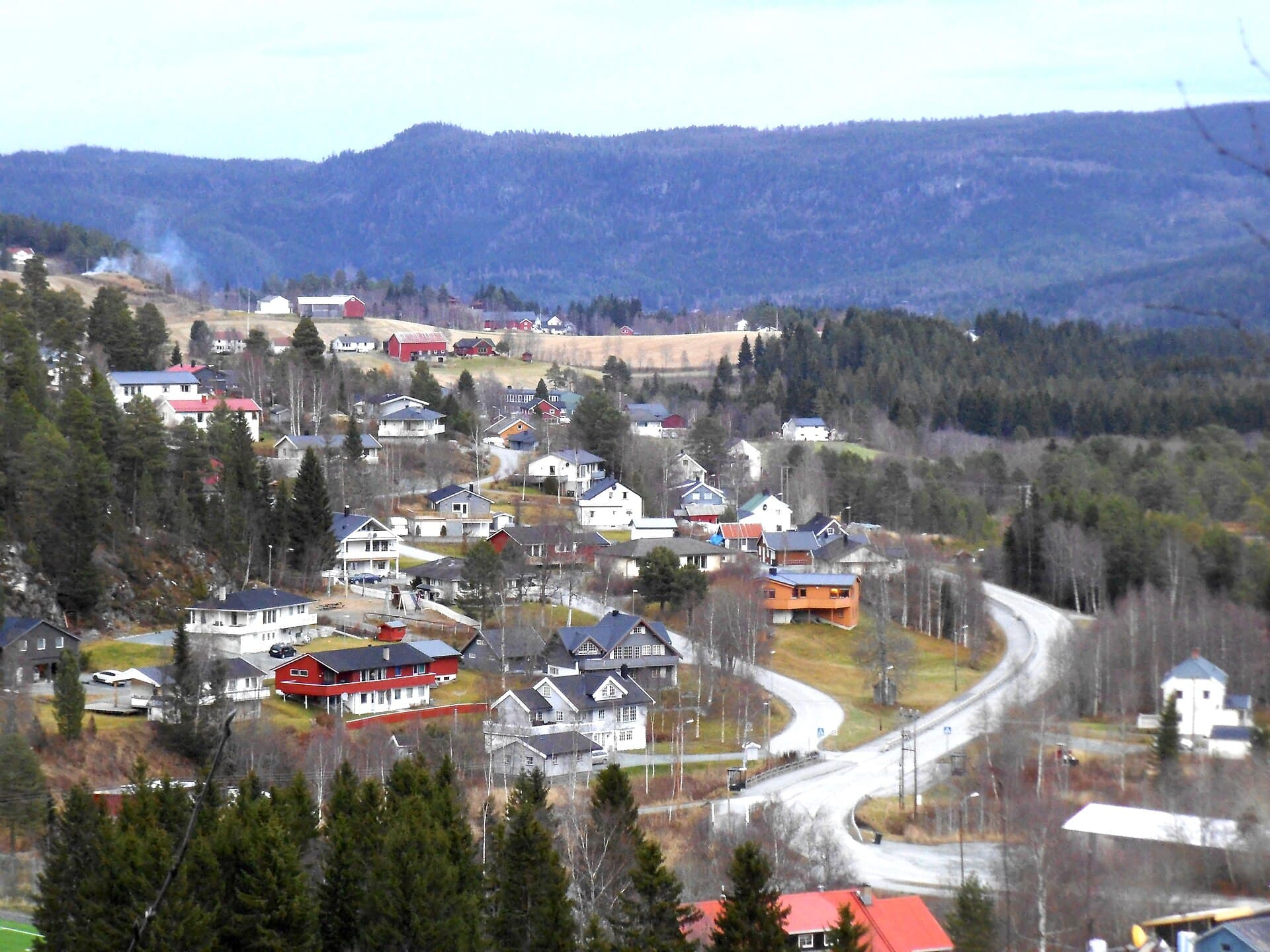 Boligfelt i Trøndelag med eneboliger langs svingete veger, jordbrukslandskap og skogkledde åser bak.