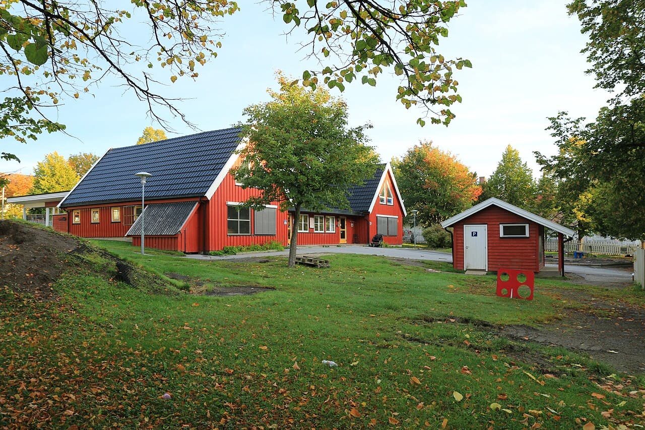 Rødmalt barnehagebygg med mørkt tak og plen med høstløv. Illustrasjonsfoto.