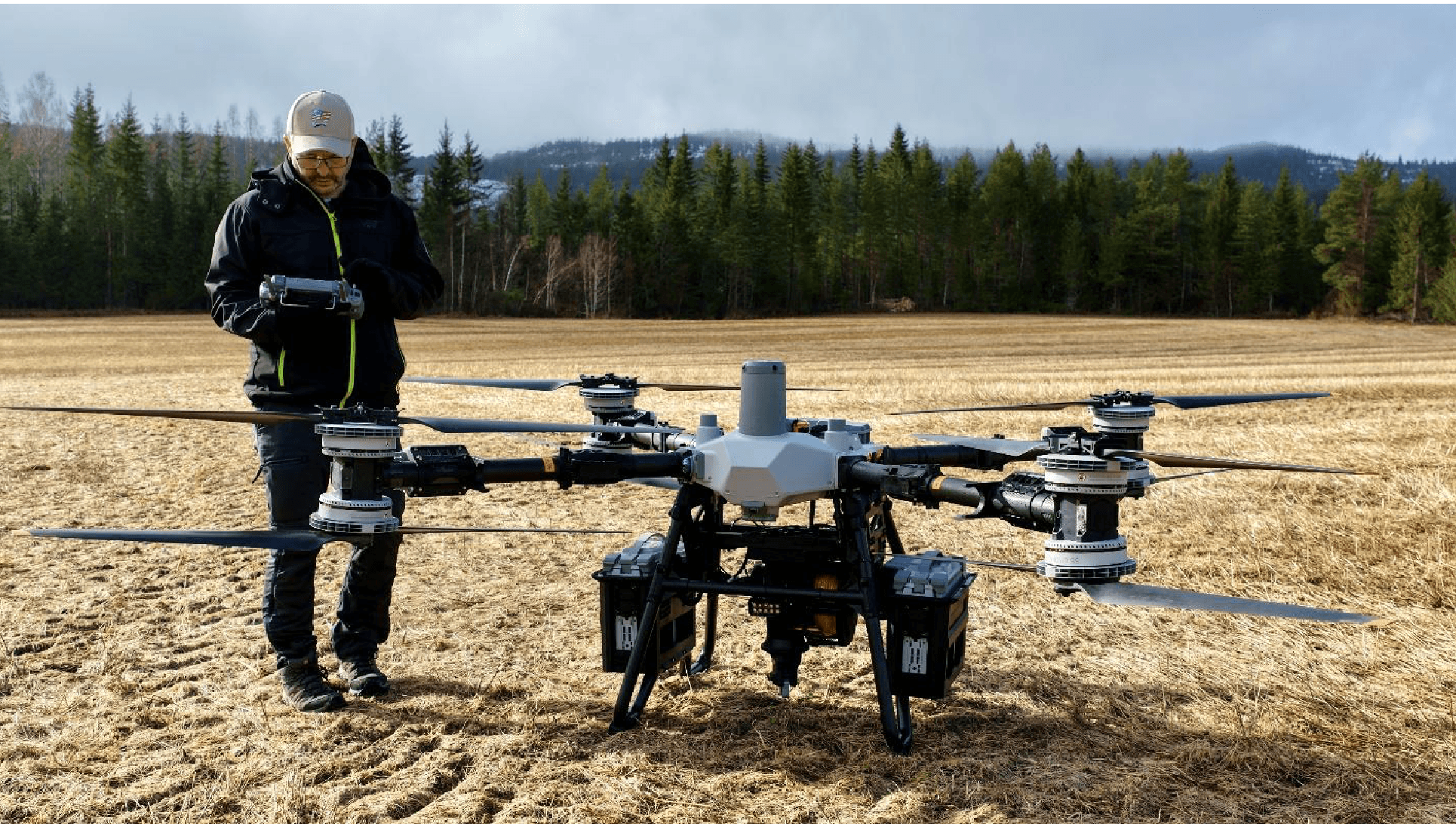 Droneoperatør står med fjernkontroll ved siden av en stor åttemotors drone på et stubbjorde, med granskog og tåkelagt ås bak.