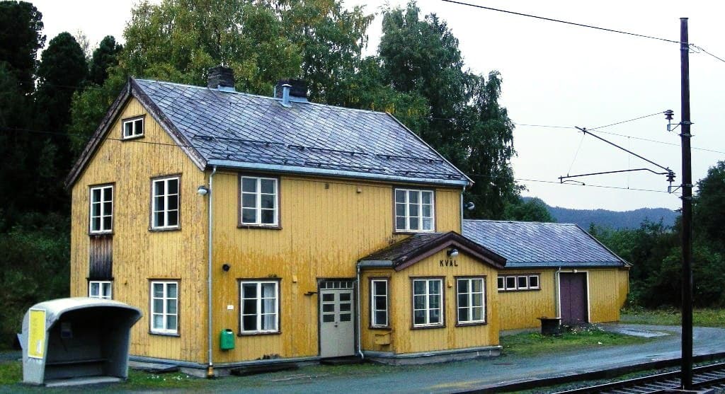 Kvål stasjon, en gul trebygning ved den elektrifiserte Dovrebanen i Melhus, med jernbanespor og skogkledde åser i bakgrunnen.