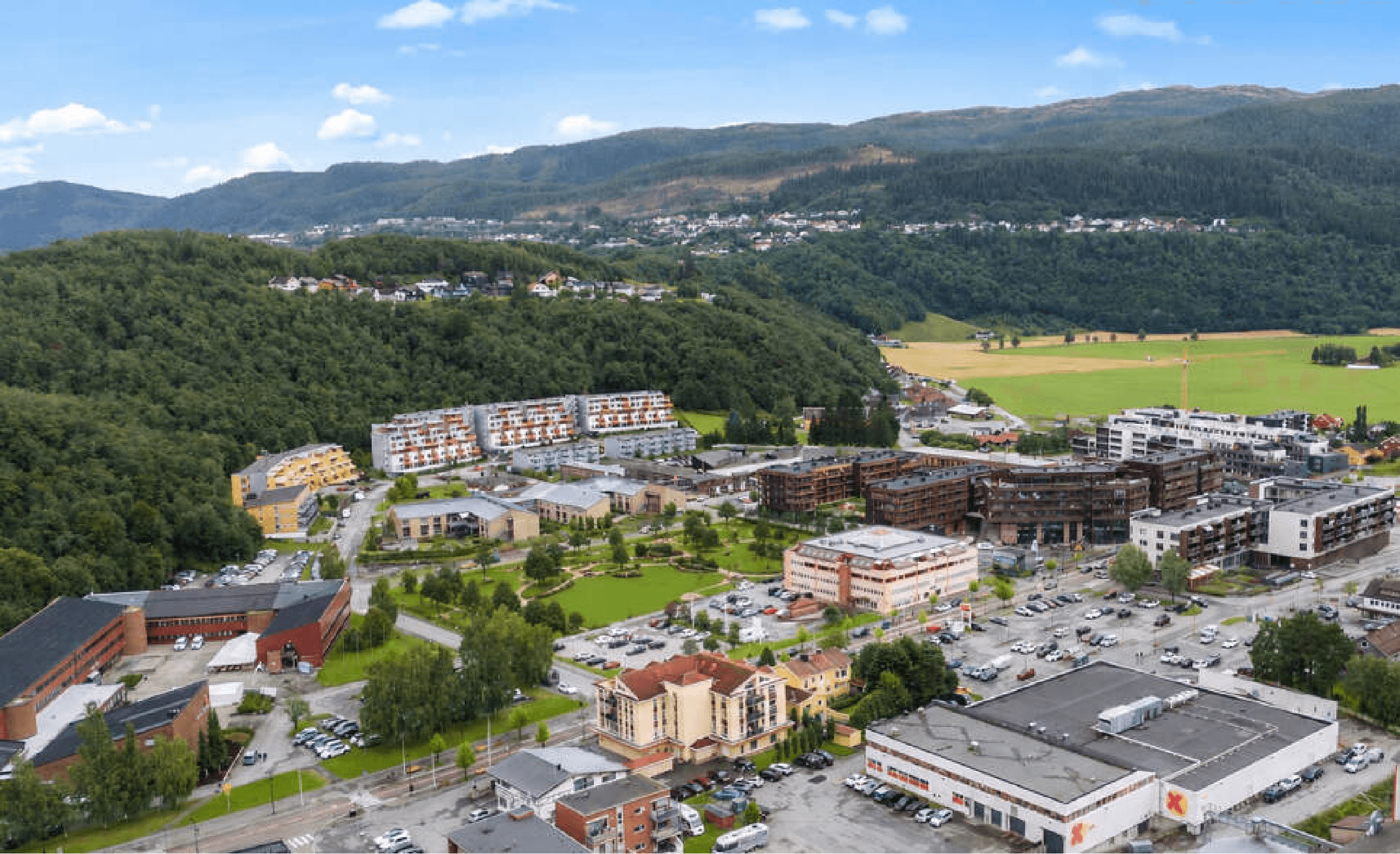 Flyfoto av Melhus sentrum med leilighetsbygg, handelsområde og grønne åser i bakgrunnen.